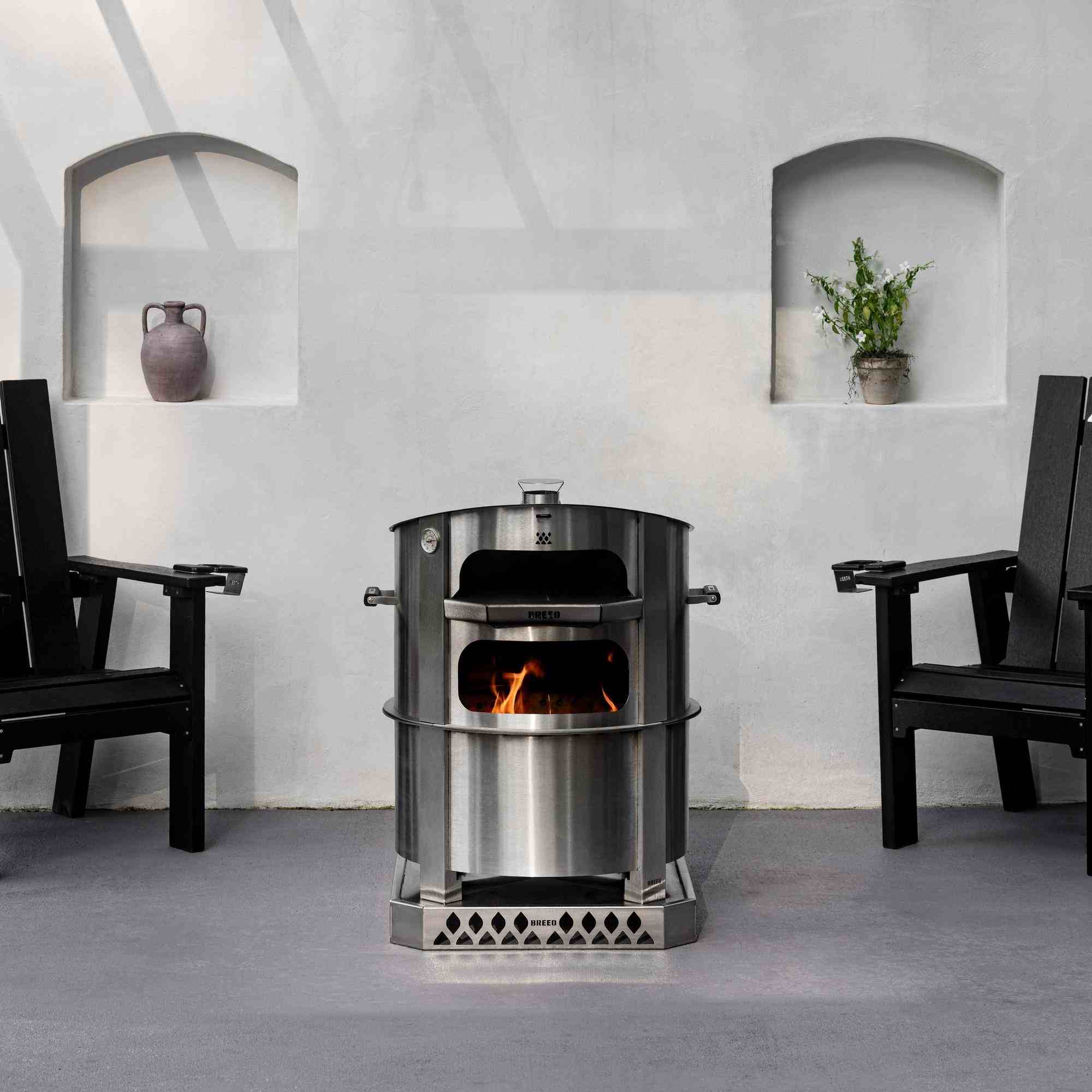 An image of the Breeo X24 stainless firepit with a Live-fire Pizza Oven and two Breeo chairs set up beside it.