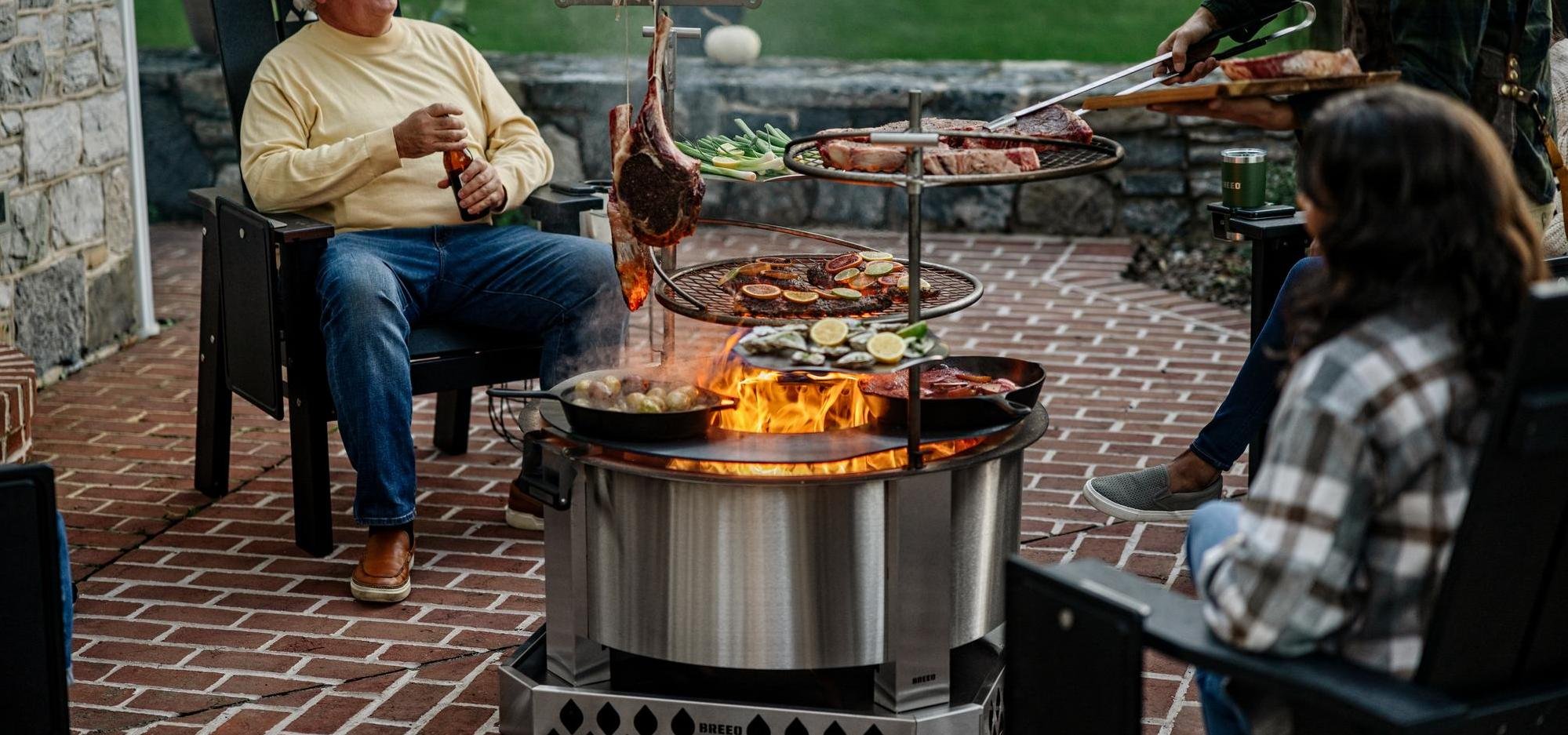 An X30 Stainless firepit set up on a patio with people sitting around it and grilling on the attached accessories including: a Searplate Griddle, 2 Outpost Grills, and a Kettle Hook