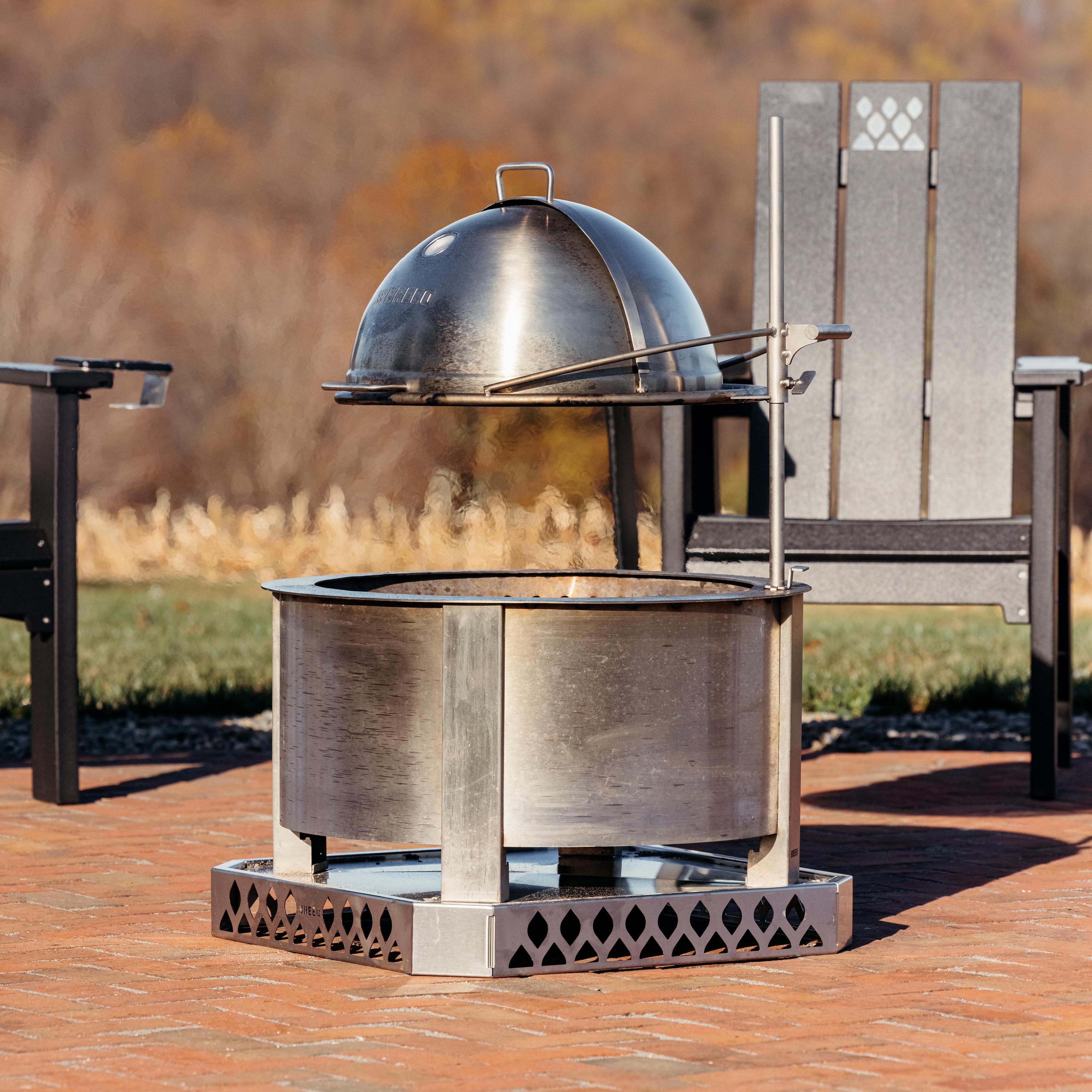 Breeo stainless steel pizza oven dome mounted on Outpost arm above Breeo fire pit, shown outdoors with X Series chair