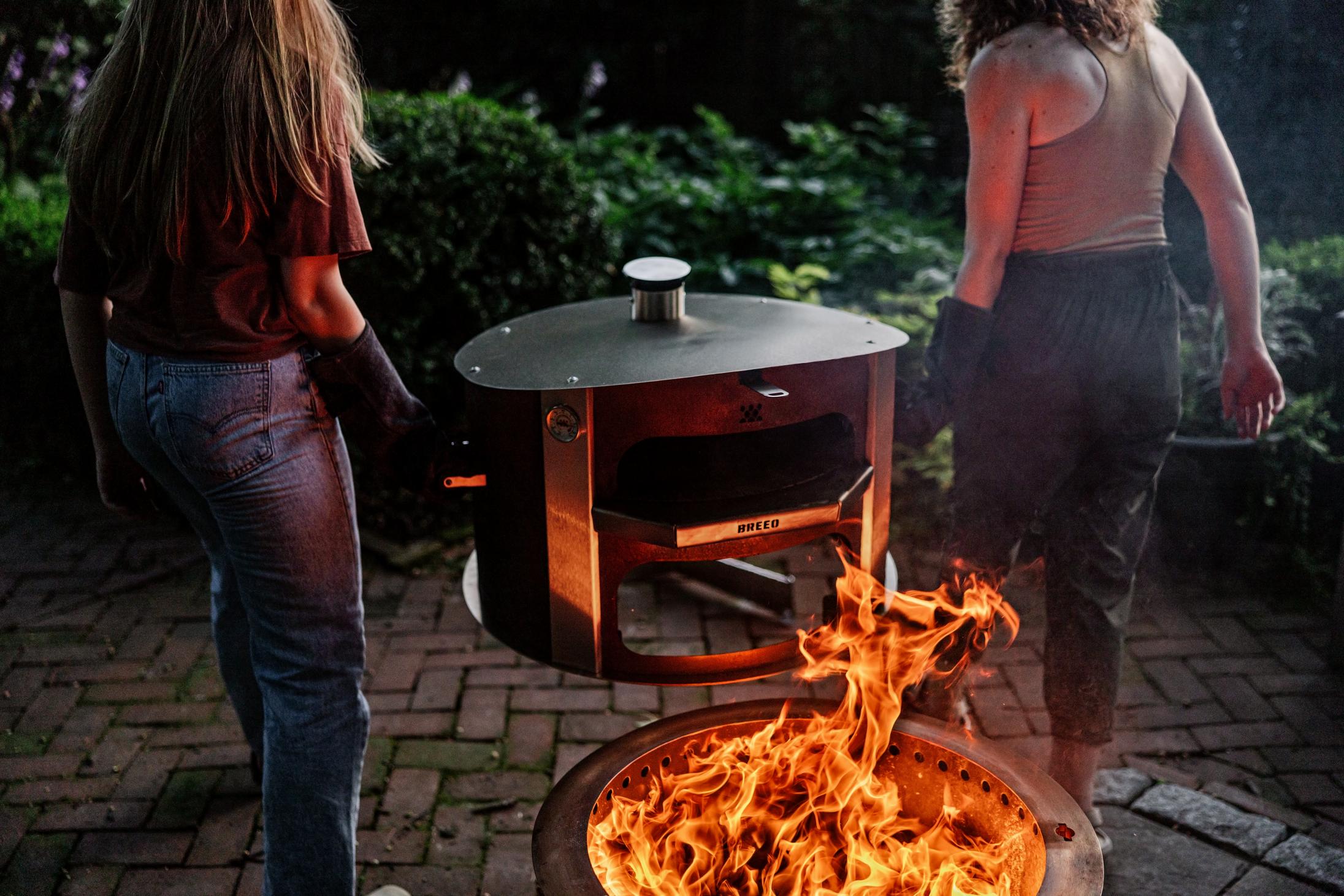 This is an image of two people removing the Breeo Pizza oven from the firepit