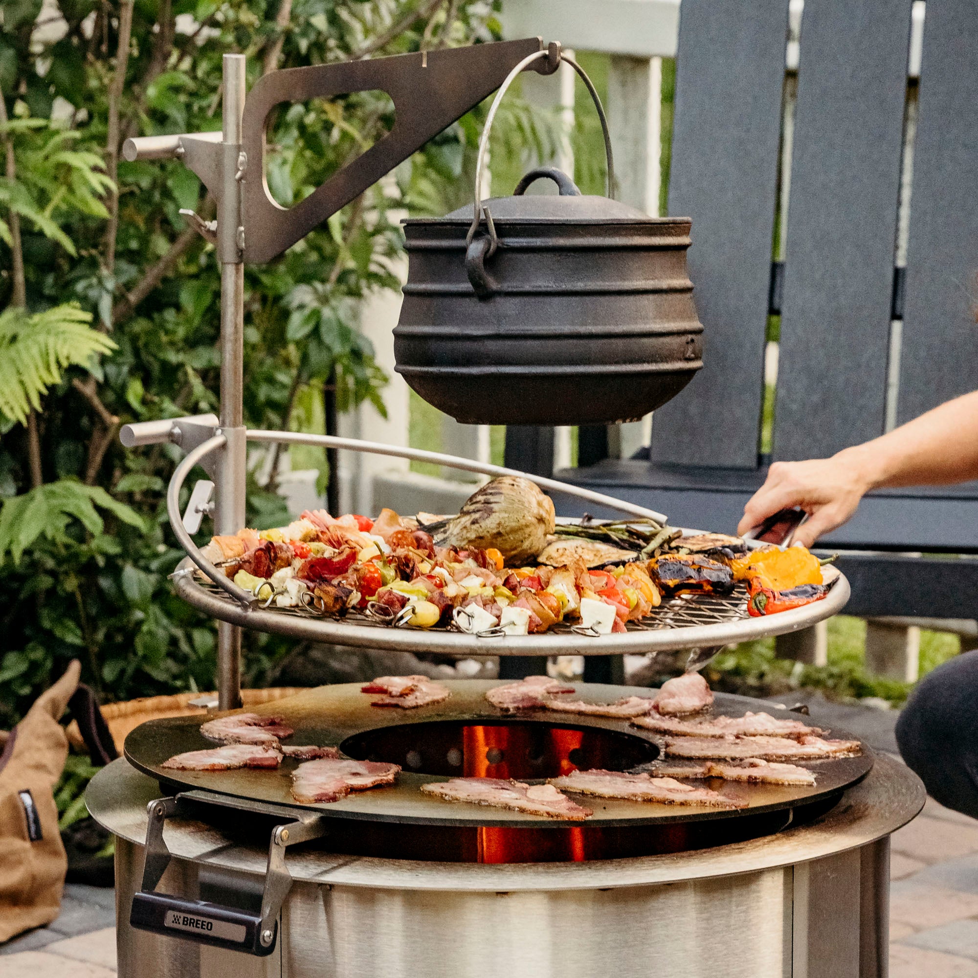 Person cooking food over breeo smokeless firepit with kettle and breeo chair in the background in outdoor setting