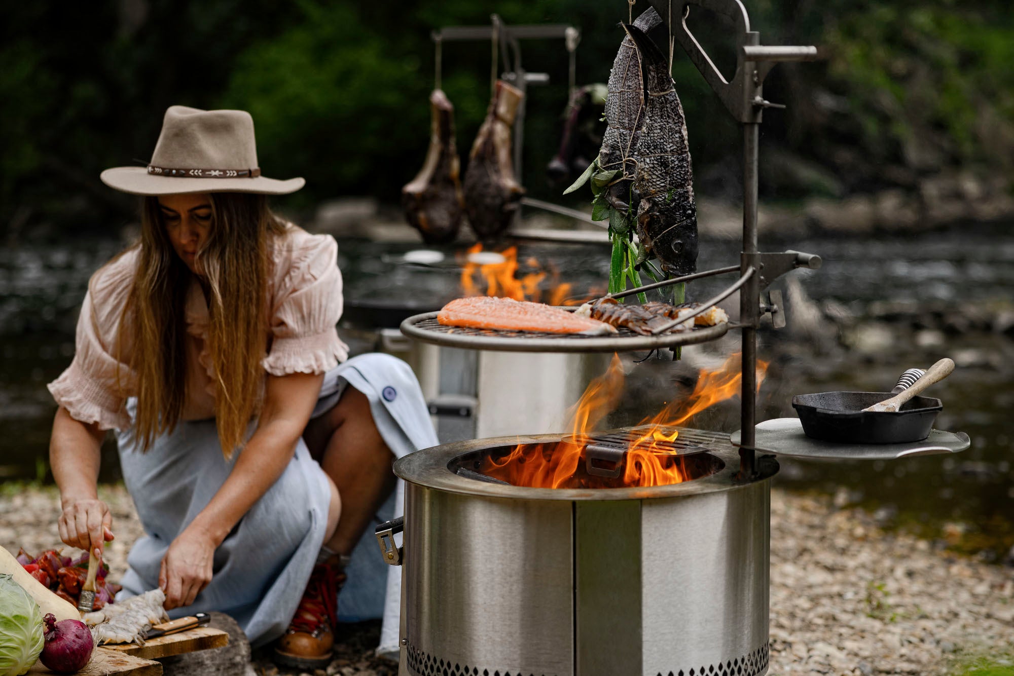 Person cooking outdoors by a breeo fire pit with various food items on skewers.