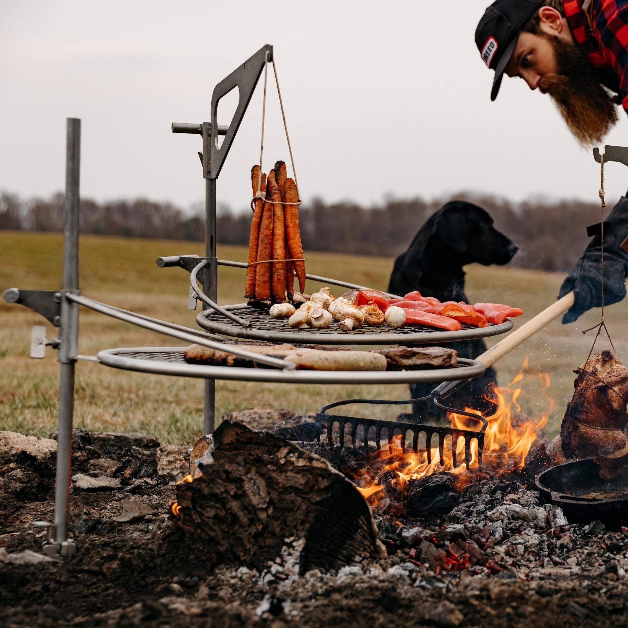This is a picture of a man raking coals while grilling over Outpost Grills with Kettle Hook Attachments