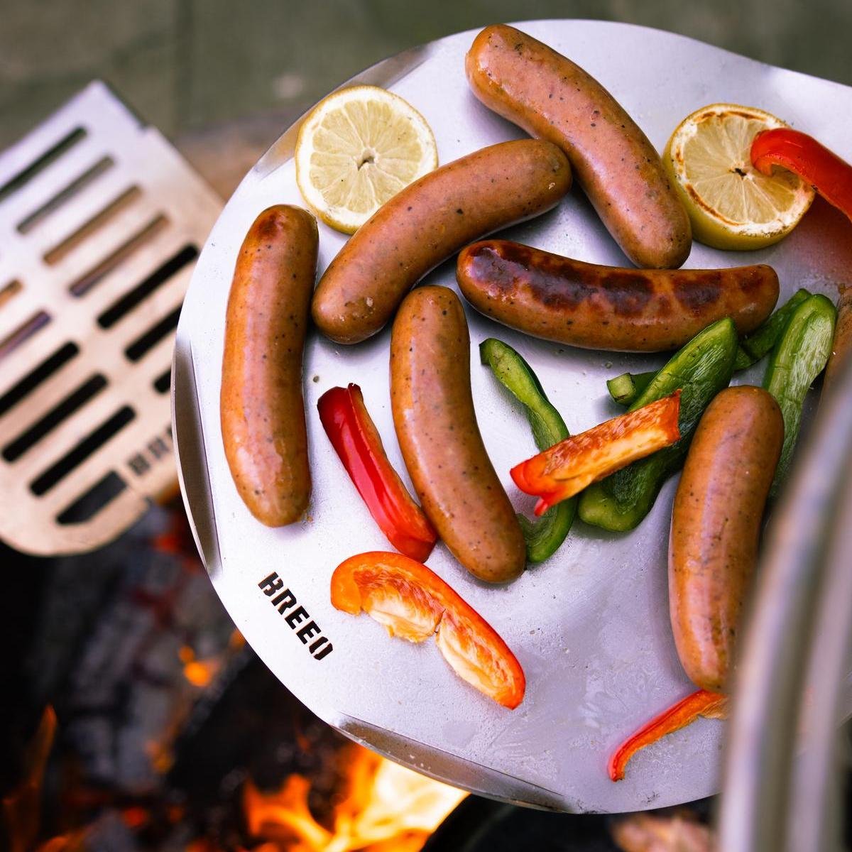 An image of grilled veggis and sausage cooking over the fire on an Outpost Sear DIsc