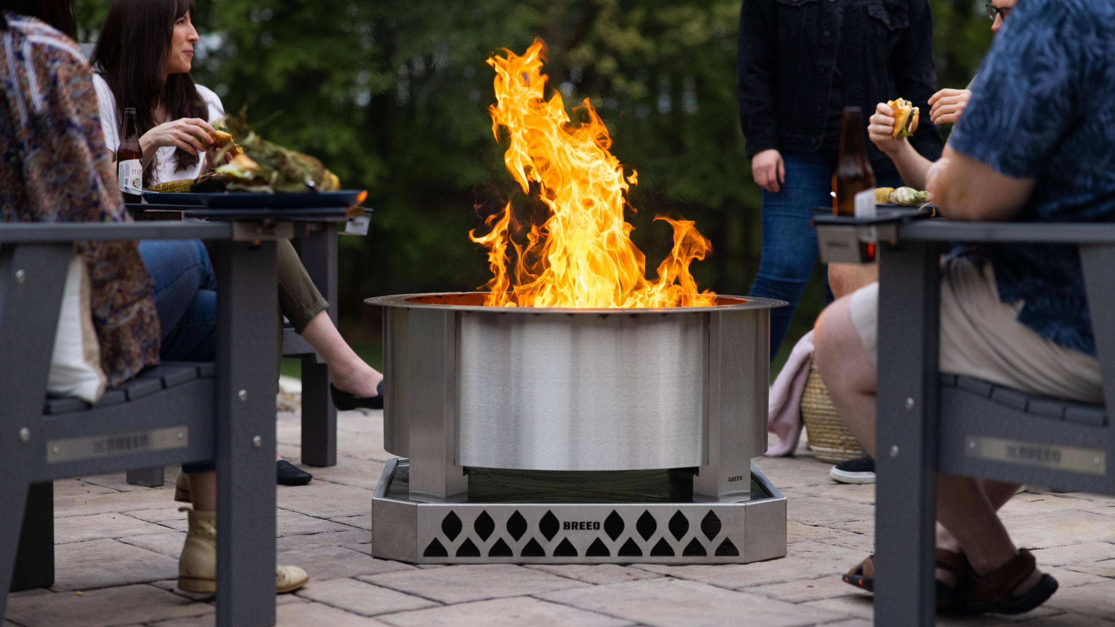 Friends gathered around a Breeo X Series fire pit and X Series chairs, enjoying grilled food and drinks on a backyard patio