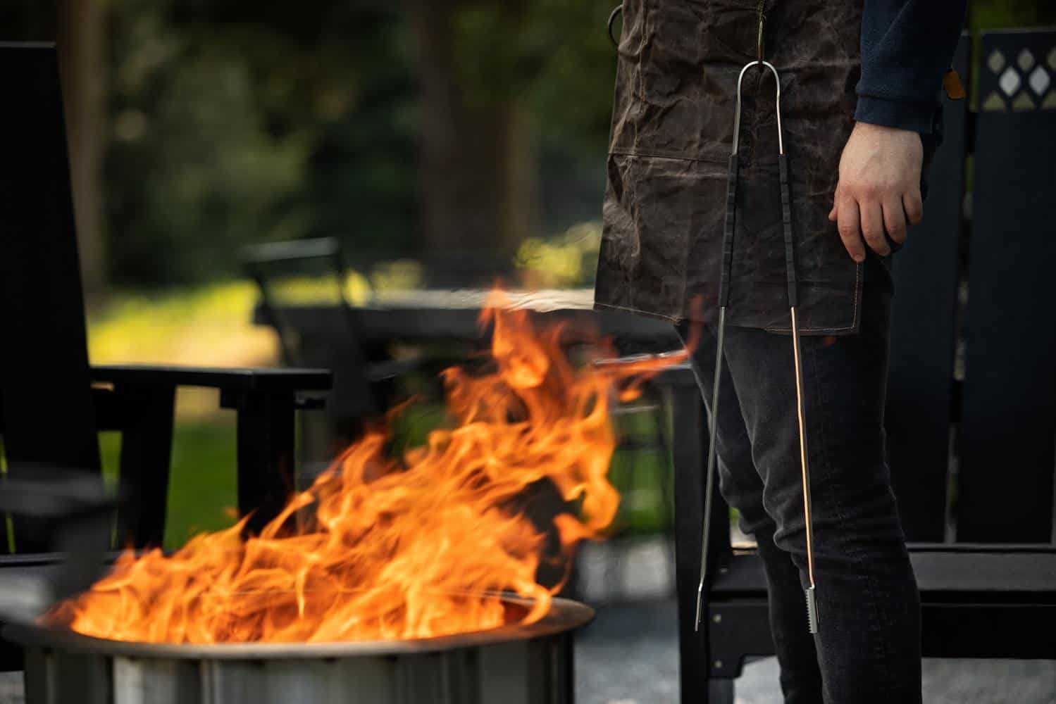 A man standing next to the fire pit wearing a canvas apron with Breeo Tongs attached to the apron