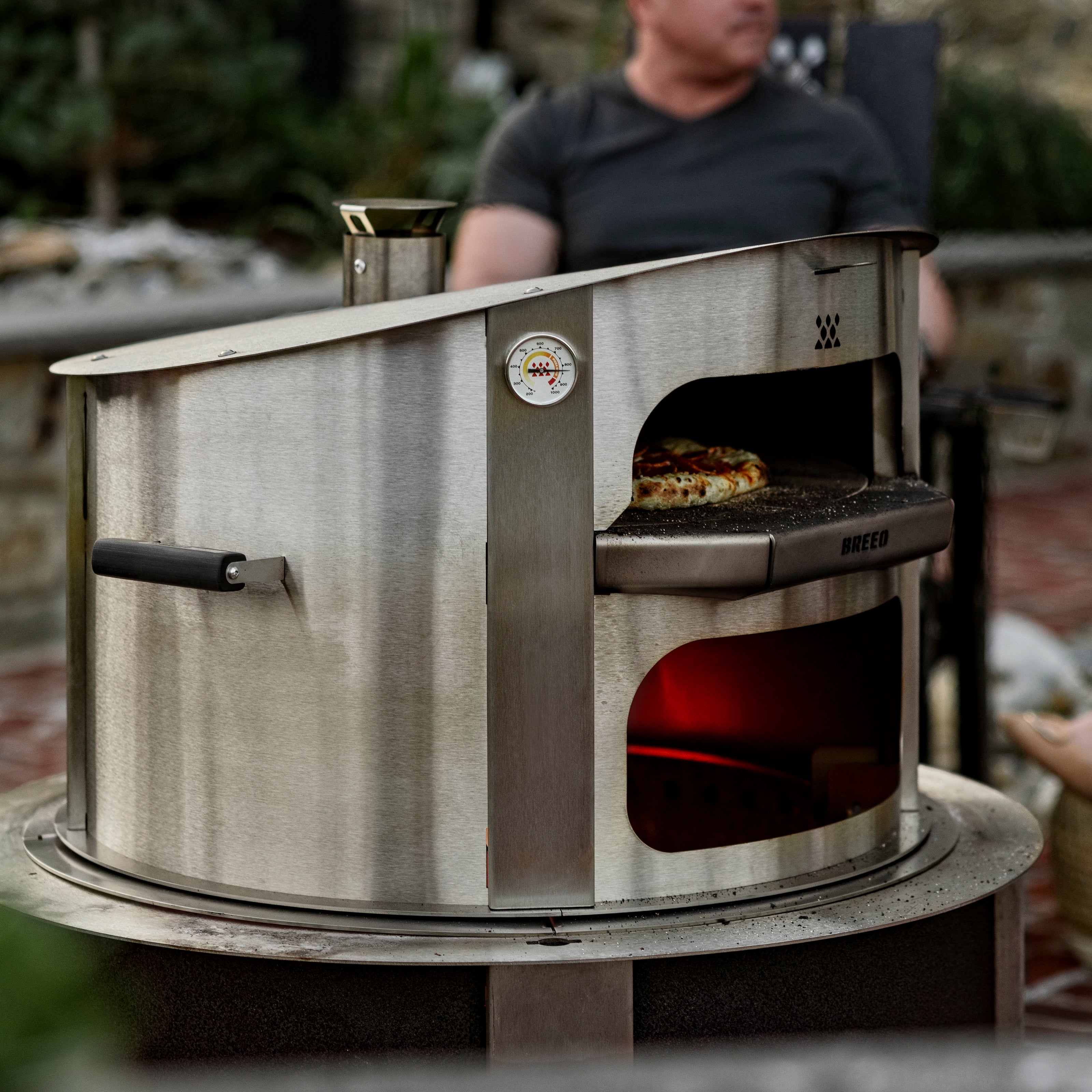 Breeo Pizza oven in outdoor setting cooking pizza with person sitting in breeo chair in background