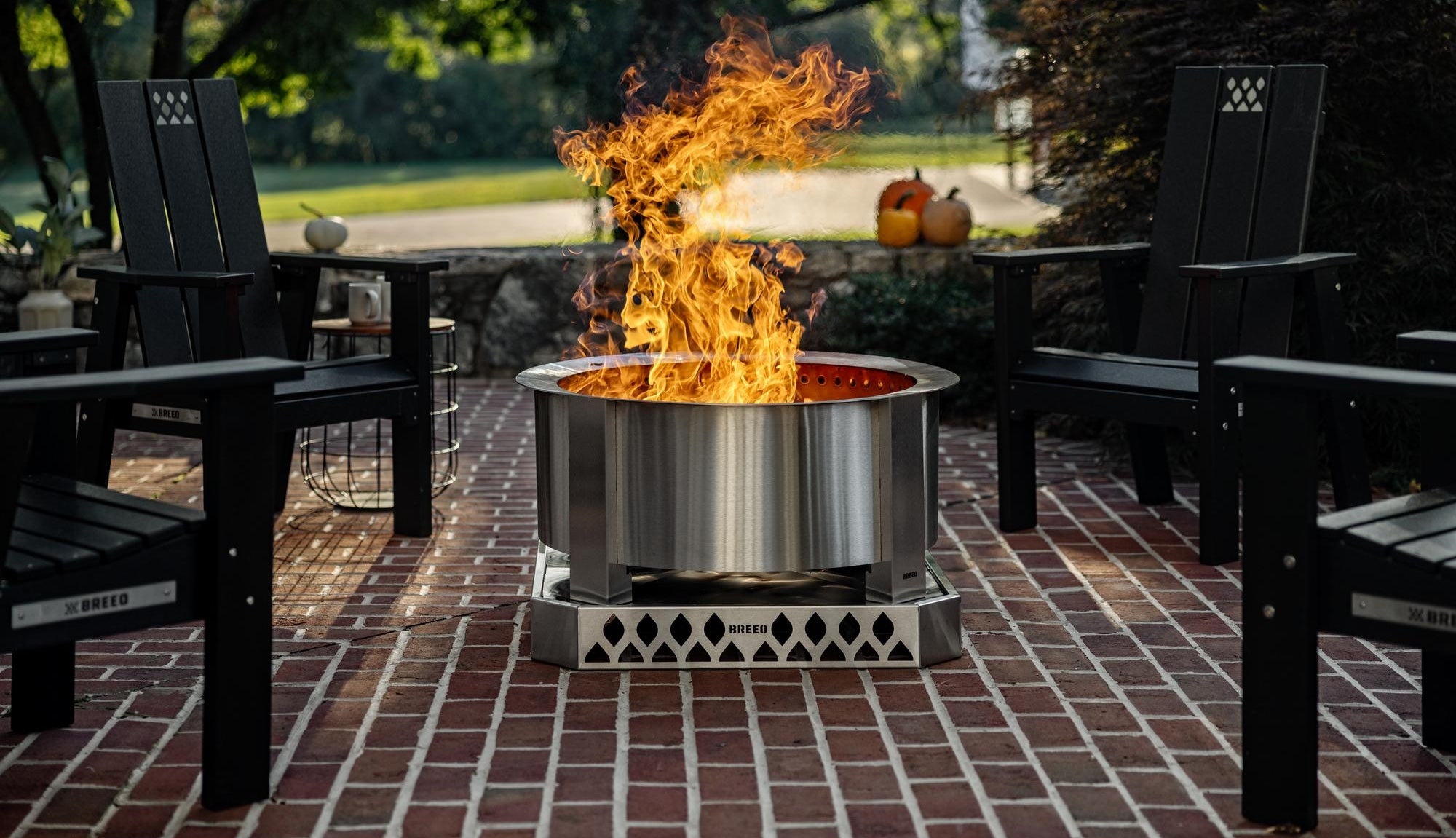 A stainless steel X 24 sits on top of a Breeo base on a brick patio surrounded by four X Series chairs.