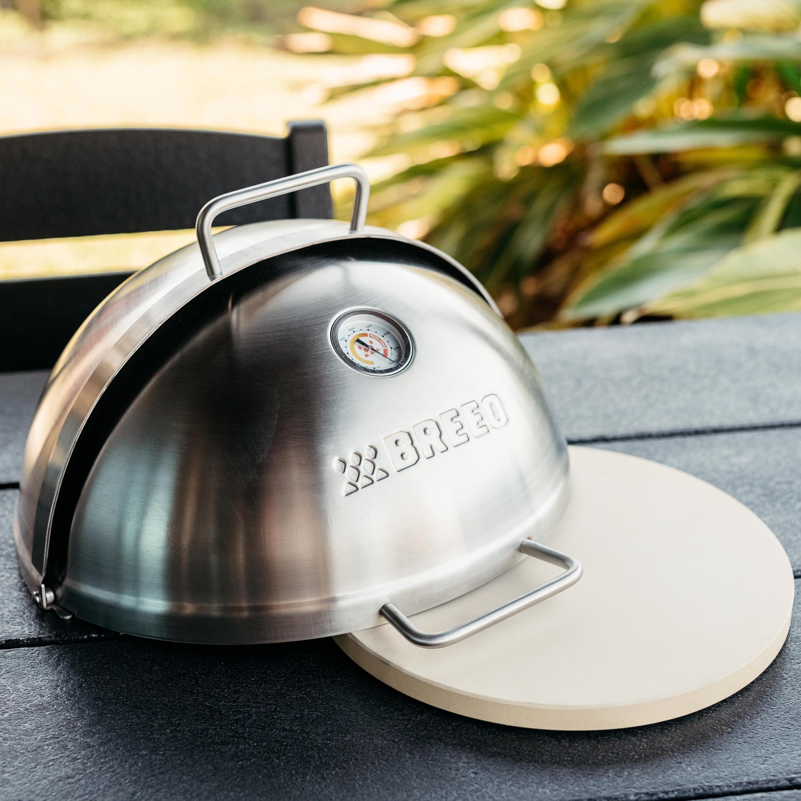 Close-up of Breeo stainless steel pizza oven dome with built-in thermometer, resting on a ceramic pizza stone atop outdoor dining table