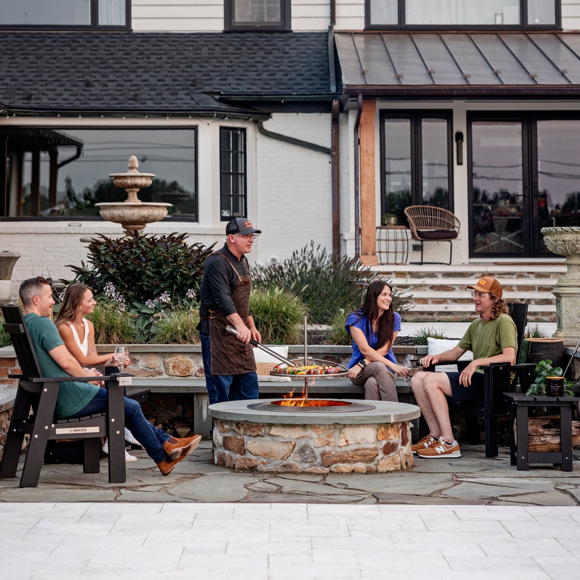 A man grilling on his patio over the Zentro fire pit with friends gathered around and sitting on X Series Furniture