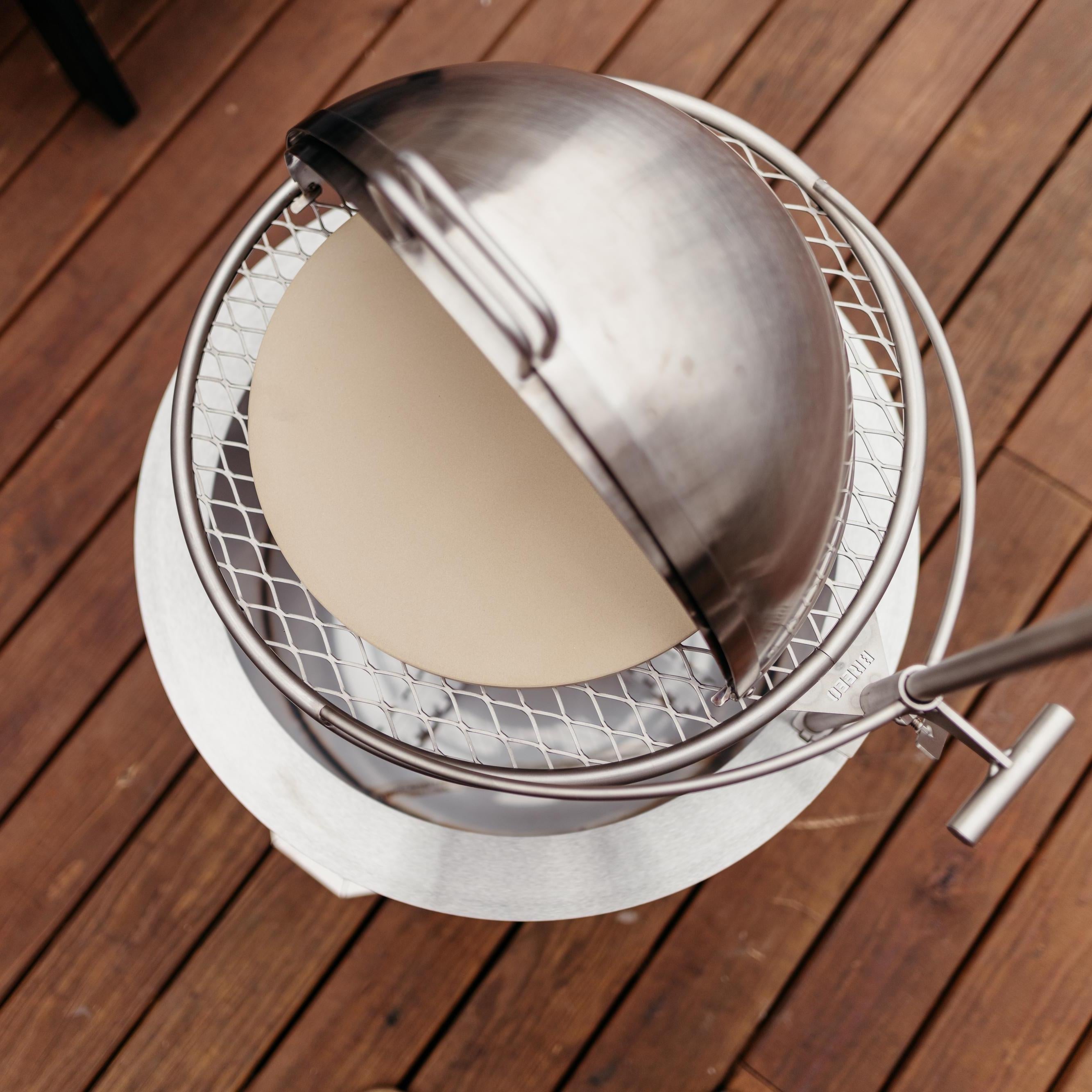 Top-down view of a Breeo live-fire grill with a ceramic pizza stone under a partially opened stainless steel dome lid, set on a wooden deck