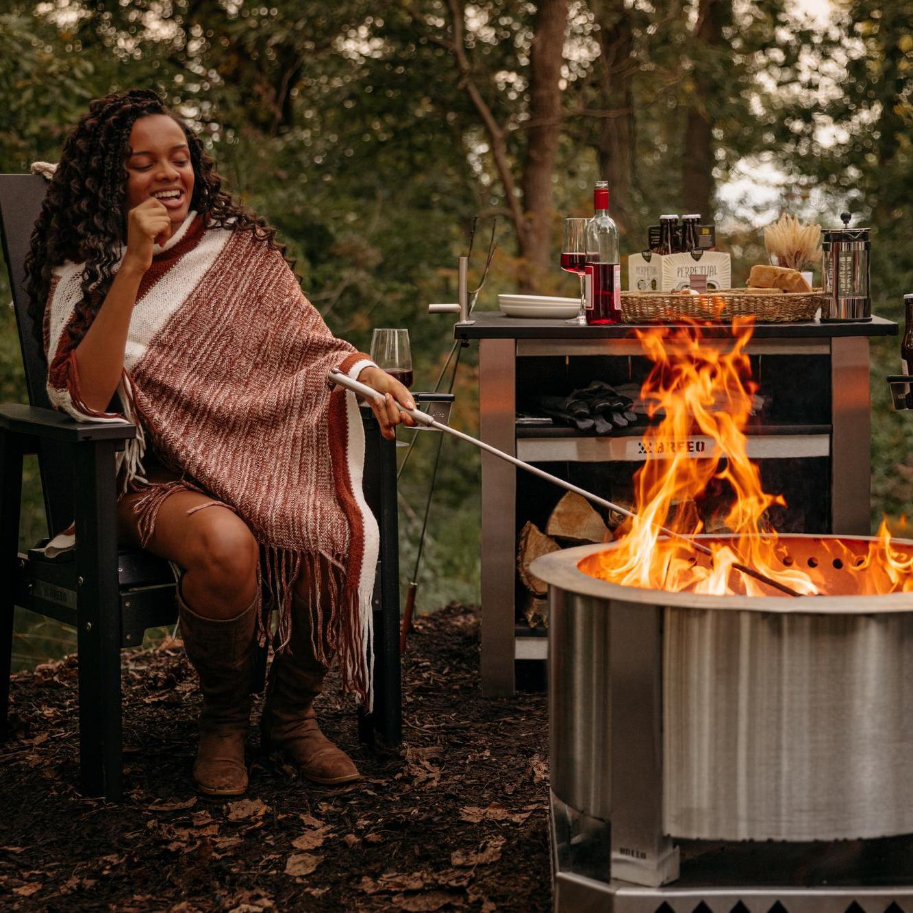 An image of a woman sitting on an X series Chair stoking the fire with a Fire Poker