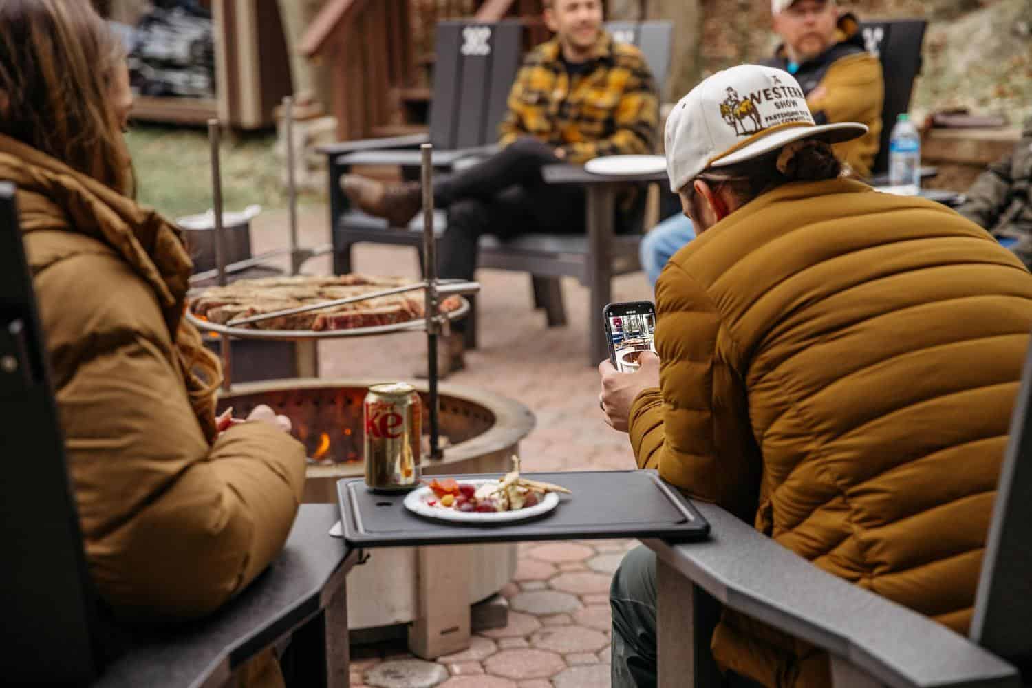 A group of people are eating around the fire on Breeo Chairs with Outrig Trays