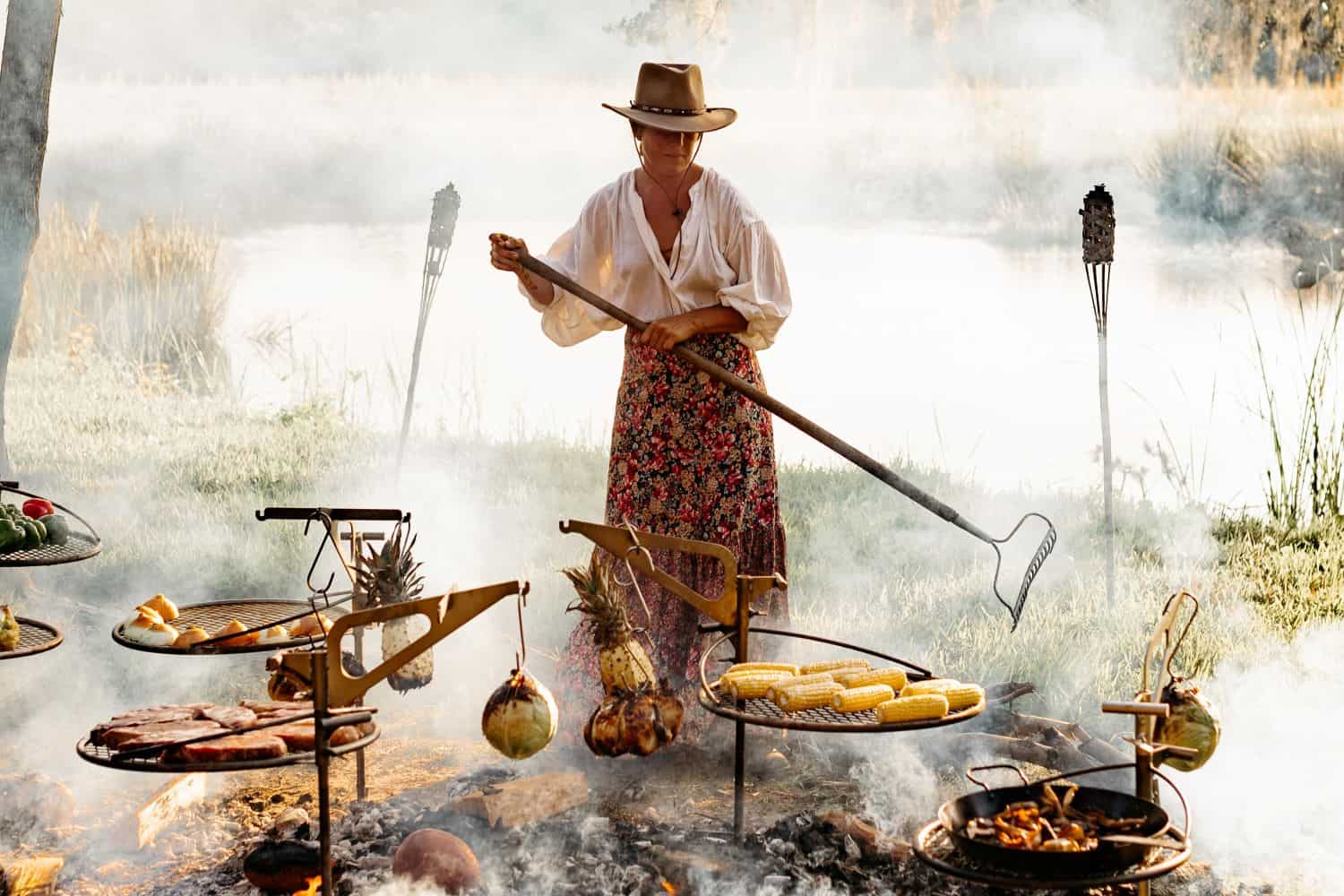 An image of a woman grilling a luxurious meal over the fire using multiple Outpost Grills and Kettle Hooks