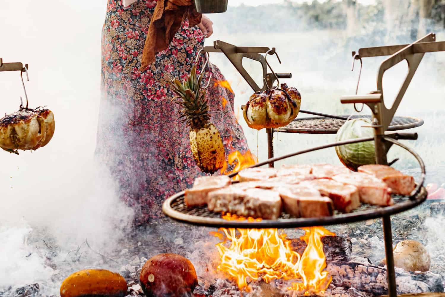 An image of a woman grilling a luxurious meal over the fire using multiple Outpost Grills and Kettle Hooks