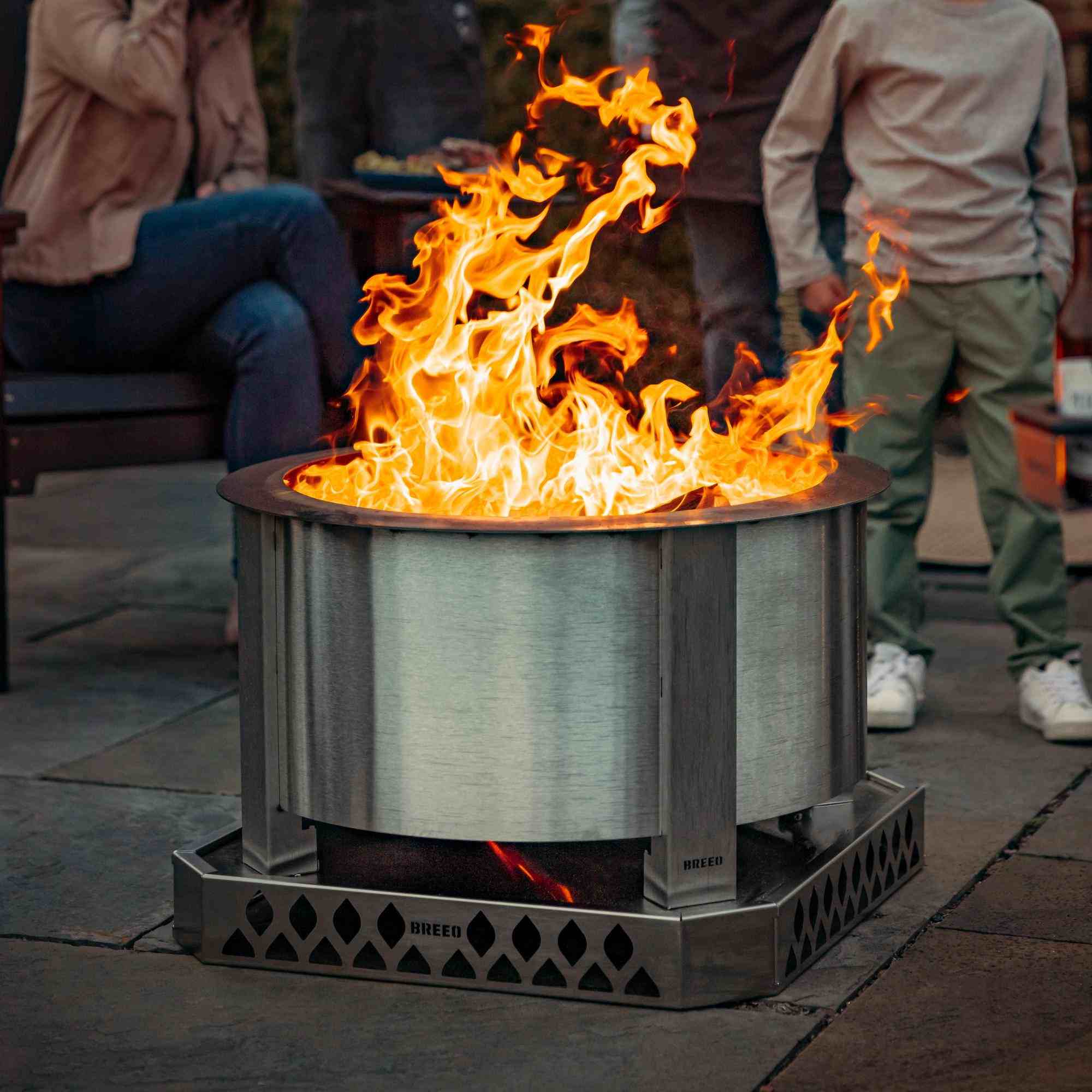 An image of the X24 Stainless firepit with a roaring fire, resting on a Breeo Base with people in the background