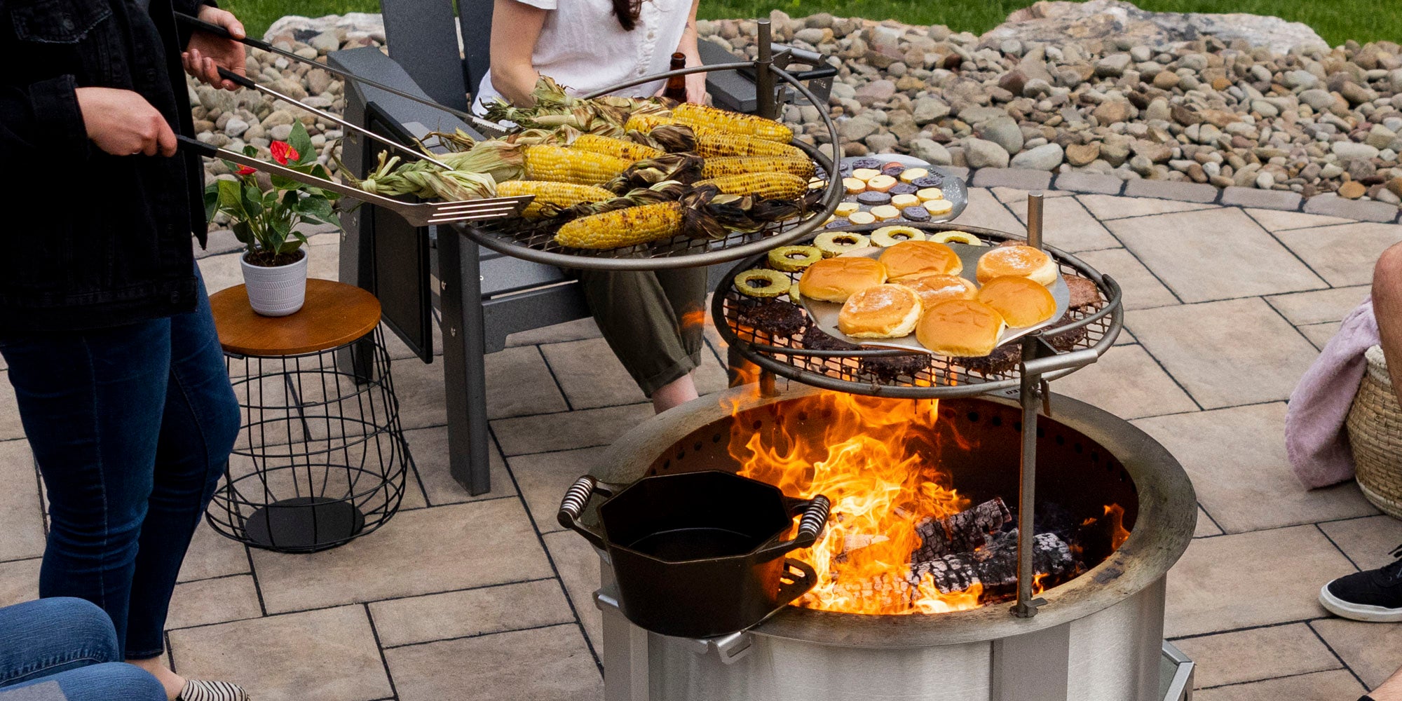 people on patio cooking food over tiered breeo smokeless firepit in outdoor setting