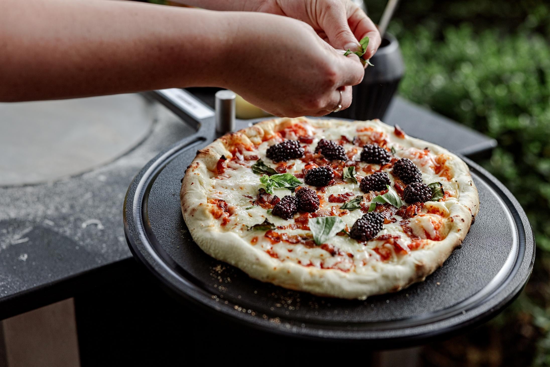 This is an image of someone sprinkling basil on a pizza resting on the Serving Board. 