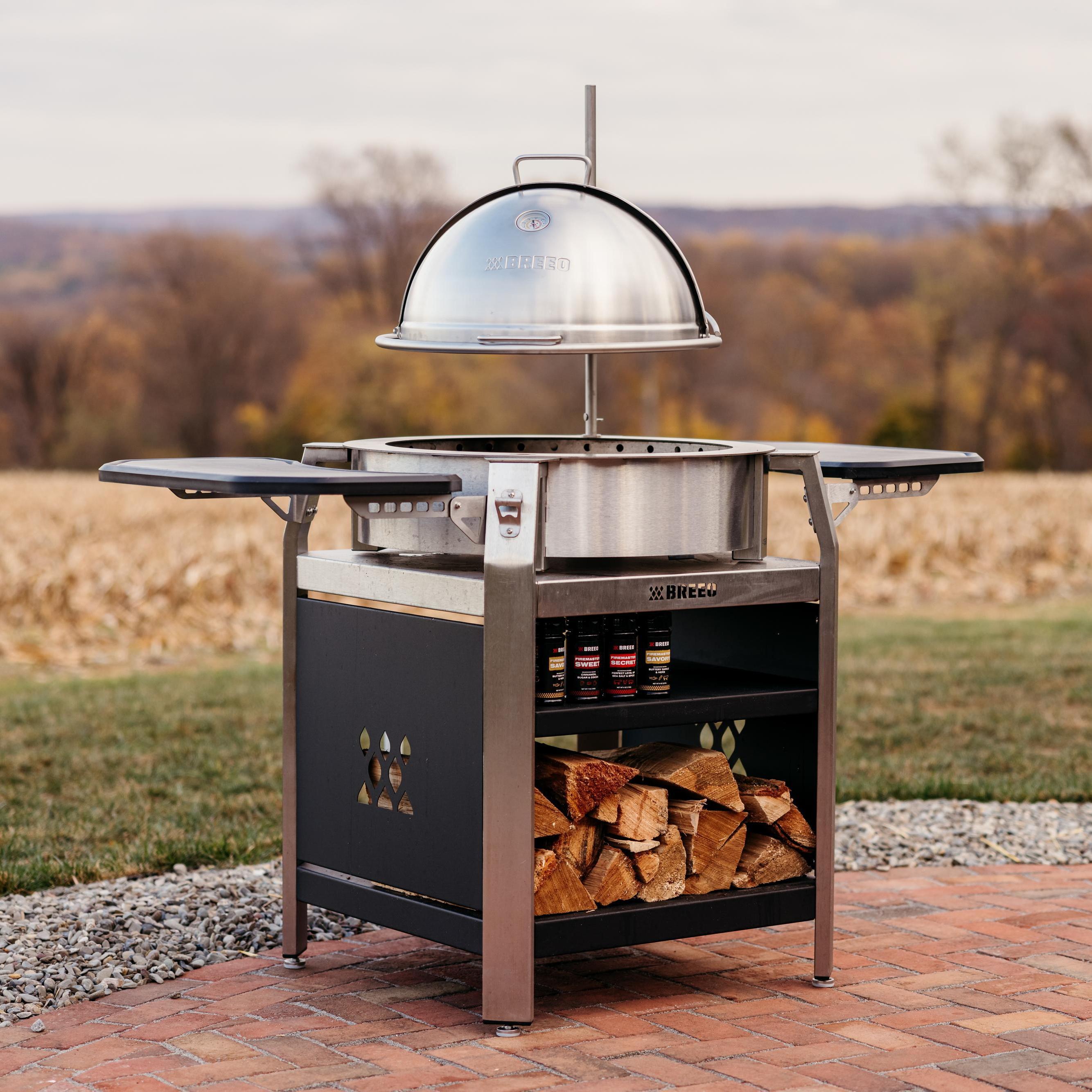 Breeo live-fire grill station with stainless steel dome lid, folding side trays, spice shelf, and wood storage, set on a brick patio overlooking a fall landscape