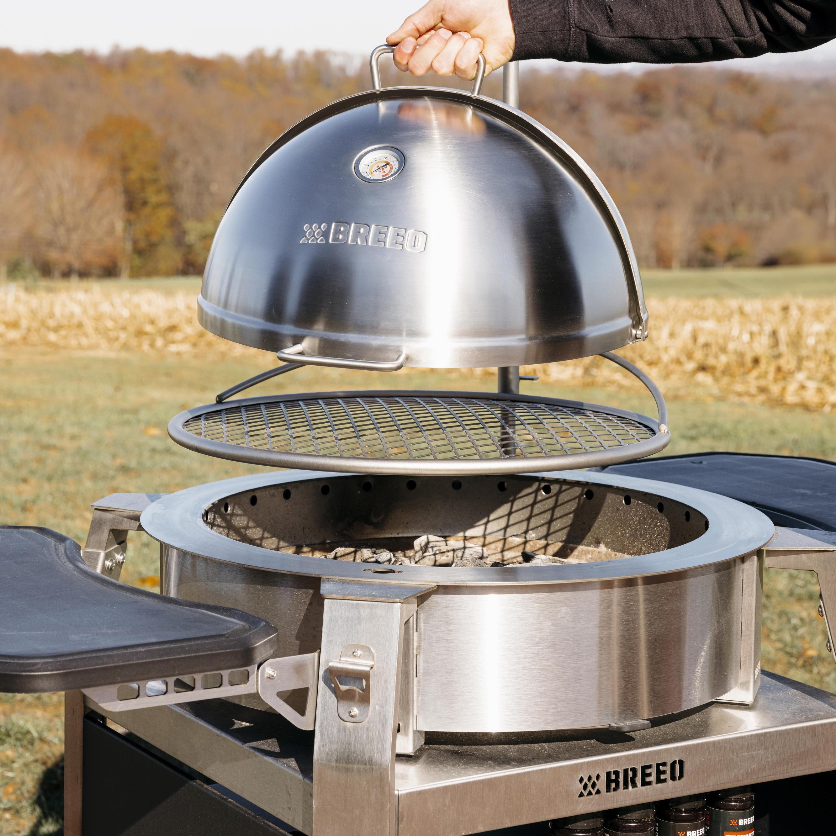 Hand lifting a stainless steel Breeo dome lid with built-in thermometer above a grill grate on a smokeless fire pit insert, set up outdoors with a scenic autumn background