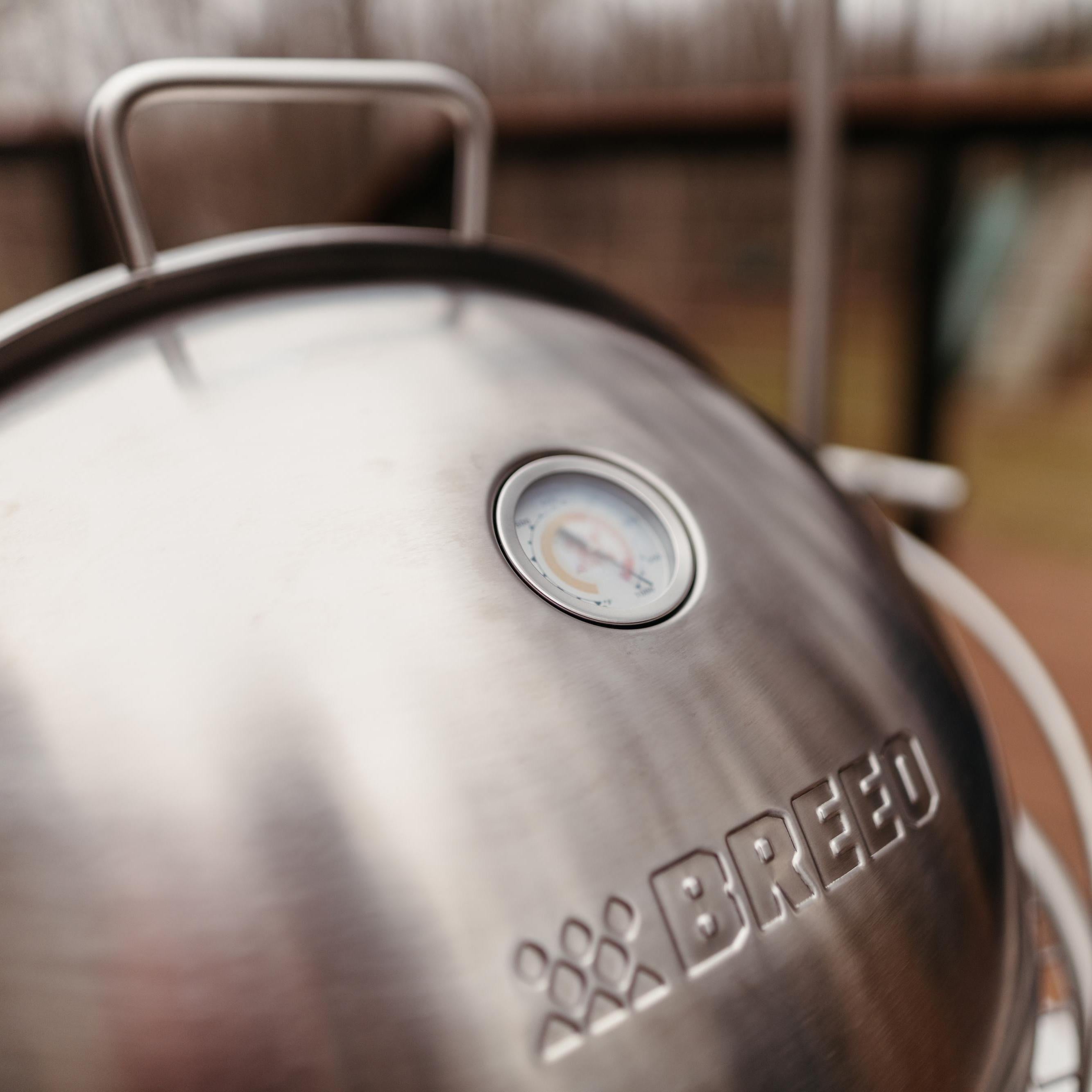 Close-up of Breeo stainless steel dome lid showing built-in temperature gauge and engraved logo, part of outdoor fire pit cooking system