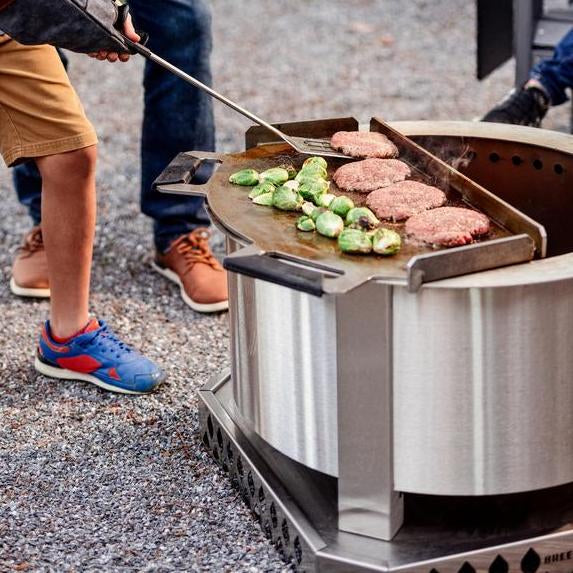 A boy getting ready to flip burgers grilling on a Flat Top attached to an X24 Stainless firepit 