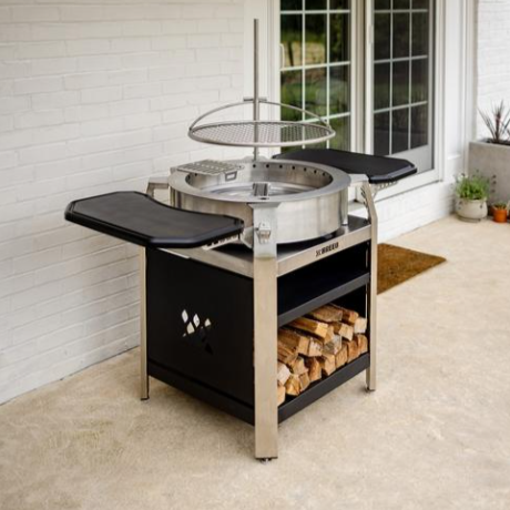 Outdoor fire pit grill with sausages, vegetables, and a hanging pot near a pool.